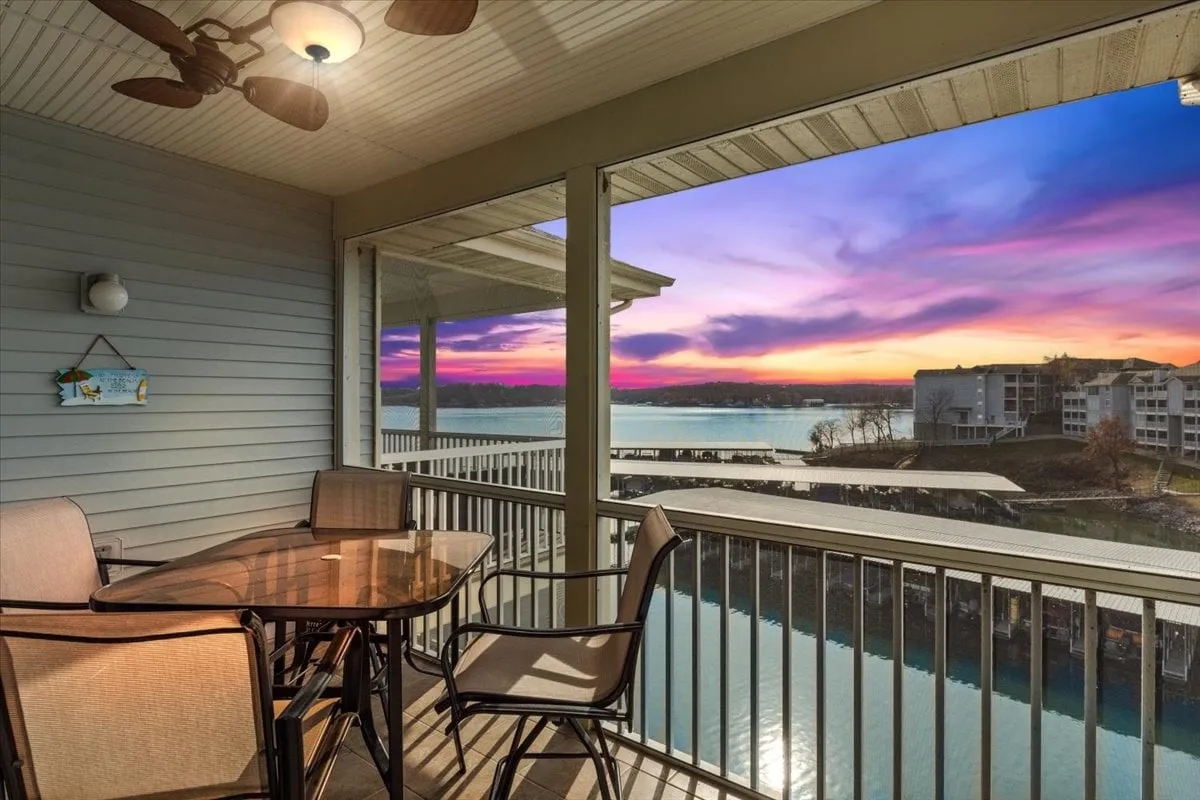 This exterior hero image captures a serene screened balcony overlooking the main channel of Lake of the Ozarks. A dining table is set on the spacious deck, perfect for gatherings, while the setting sun casts a warm glow across the tranquil water. The inviting space promises relaxation, outdoor dining, and breathtaking sunset views.