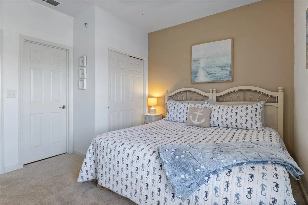 A cozy bedroom with nautical-themed bedding, an anchor decorative pillow, artwork above the bed, and a nightstand with a lamp. The scene is softly lit, emphasizing warmth and comfort.
