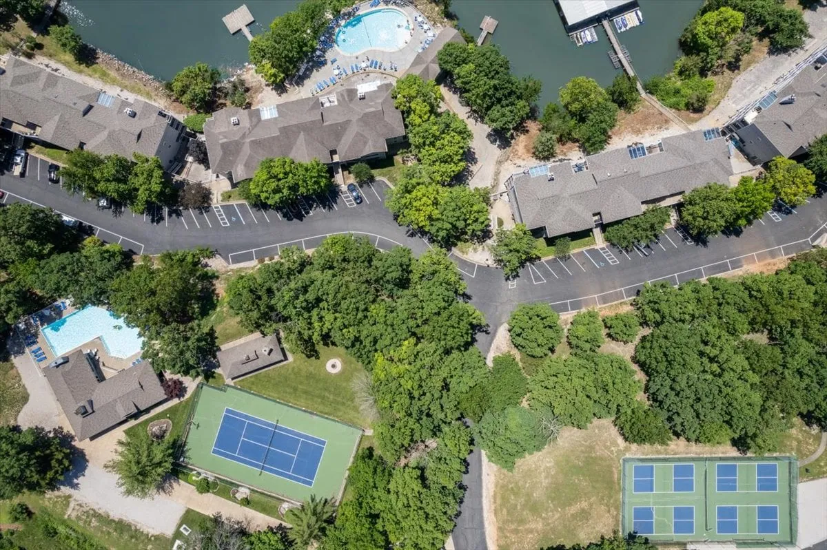 A wide view of a beautifully landscaped resort-style amenity area featuring two sparkling swimming pools, tennis courts, and lush green spaces, all nestled near the waterfront. The pools have inviting lounge chairs and tables, while the tennis courts are well-maintained and easily visible in the scene.