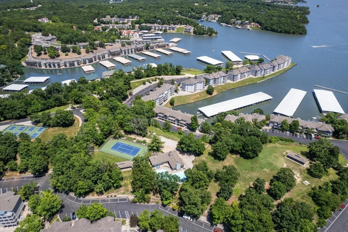A panoramic exterior shot capturing the lakeside community with tennis courts, a shimmering pool, marina, and inviting green spaces, all framed by residential buildings and the tranquil lake beyond. The area feels active, scenic, and well-integrated with nature.