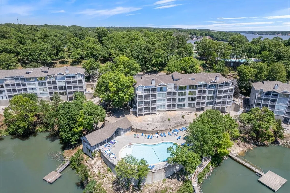 The image displays a sparkling swimming pool surrounded by lush greenery and a clear view of the waterfront and residential buildings. The inviting pool area hints at sun-filled days spent swimming, sunbathing, or gathering for a poolside barbecue with loved ones.