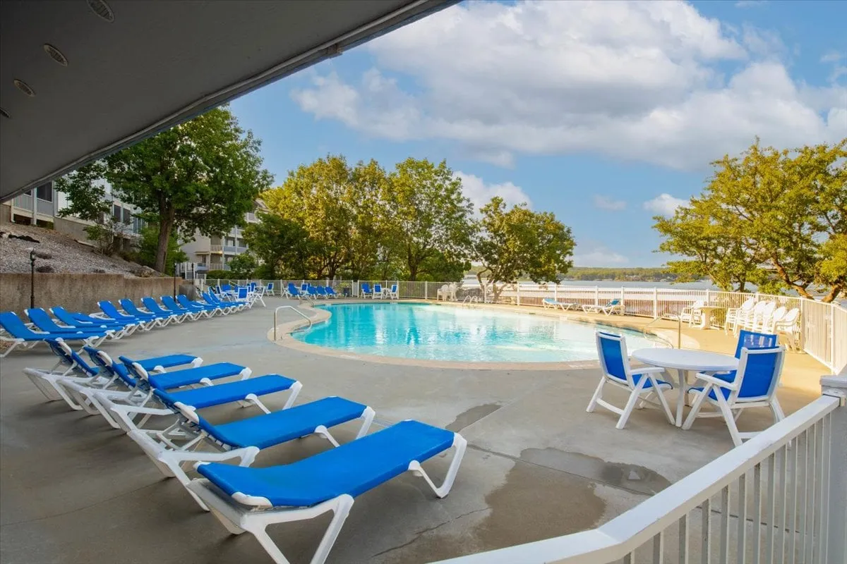 A tranquil swimming pool area framed by lounge chairs, a table and chairs for outdoor relaxation, and lush trees. Beyond the pool, a serene water view hints at the lakeside setting, offering an inviting retreat for sunbathing, swimming, or sharing peaceful moments together.