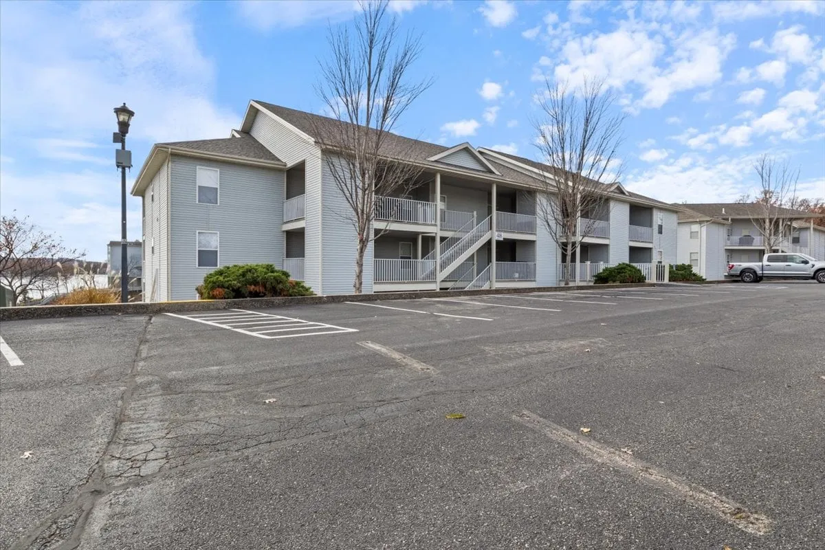 The photo shows the exterior of the apartment building with landscaped green areas, a parking lot, and people casually socializing or strolling in front. The overall atmosphere is serene and welcoming, and the image hints at easy access to local attractions and the lively community context.