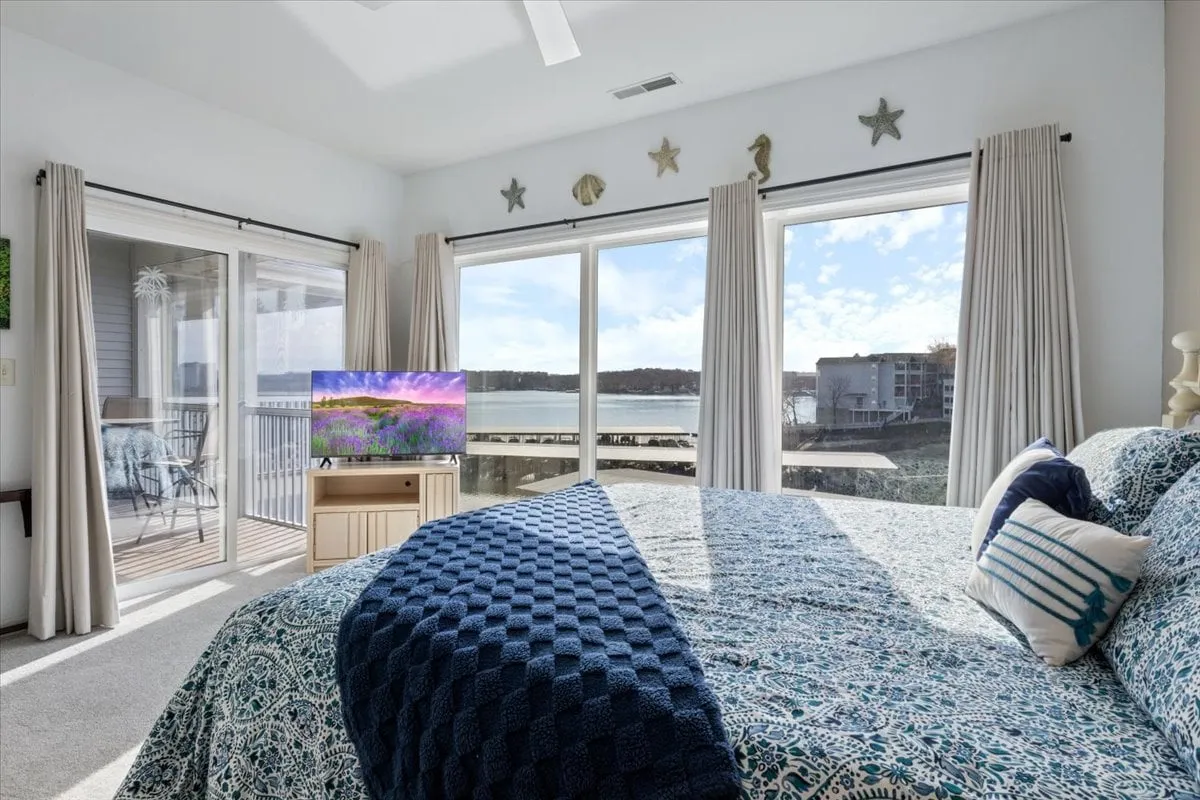 A serene bedroom with a comfortable bed adorned with decorative pillows, large windows offering a beautiful waterfront view, and a TV on the bedside table. The soft natural light and gentle colors create a peaceful, inviting atmosphere perfect for rest.