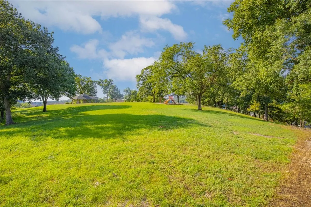 A cheerful, sunlit playground area with vibrant play equipment, a spacious canopy structure for shade, and expansive grassy lawns surrounded by mature trees. The scene feels safe, family-friendly, and perfect for active play and picnics.